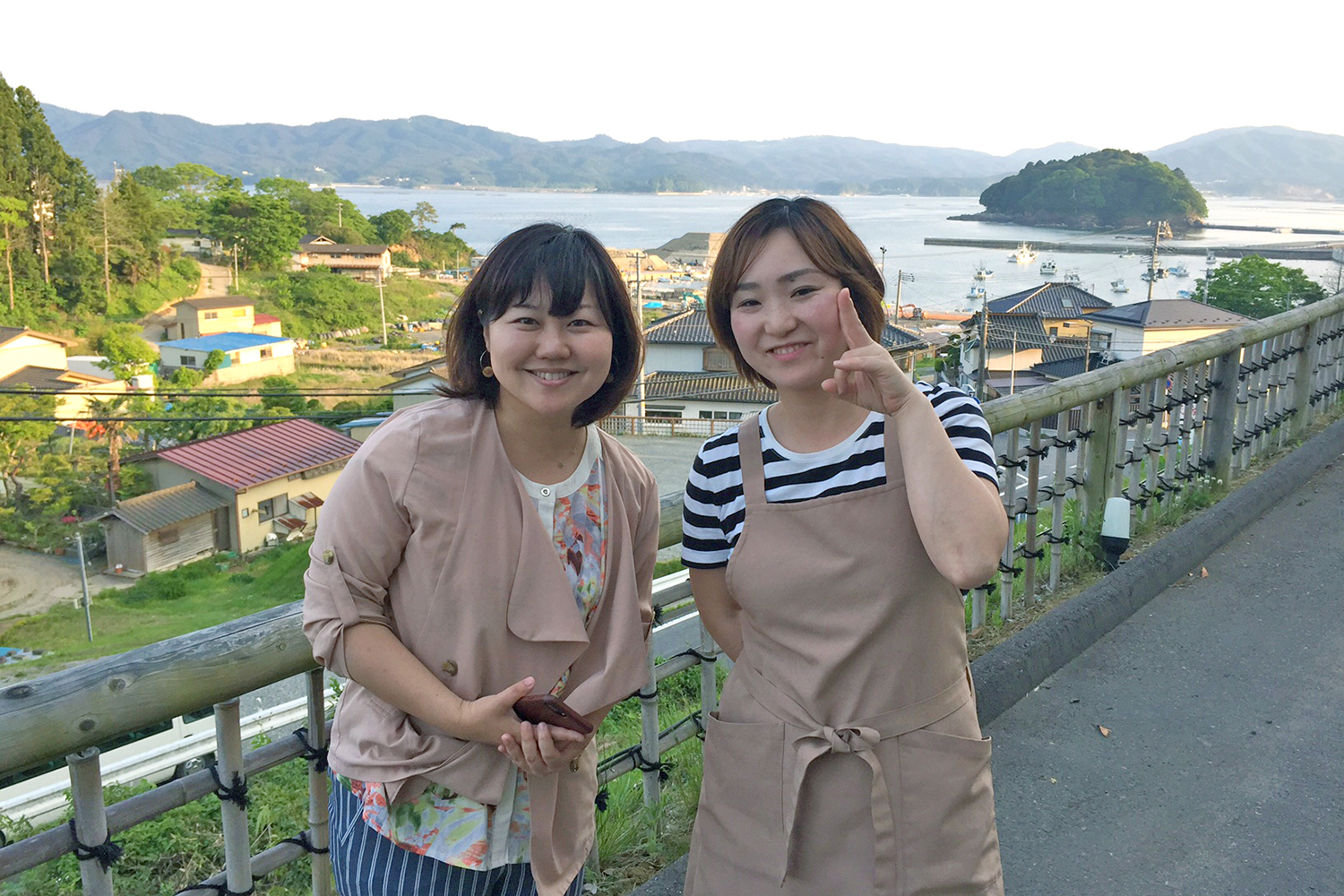 遠くに山並みが見える海辺の住宅地を見下ろす高台で、女性リポーターとベージュのエプロンをつけた女性が並んで笑顔で立っている写真