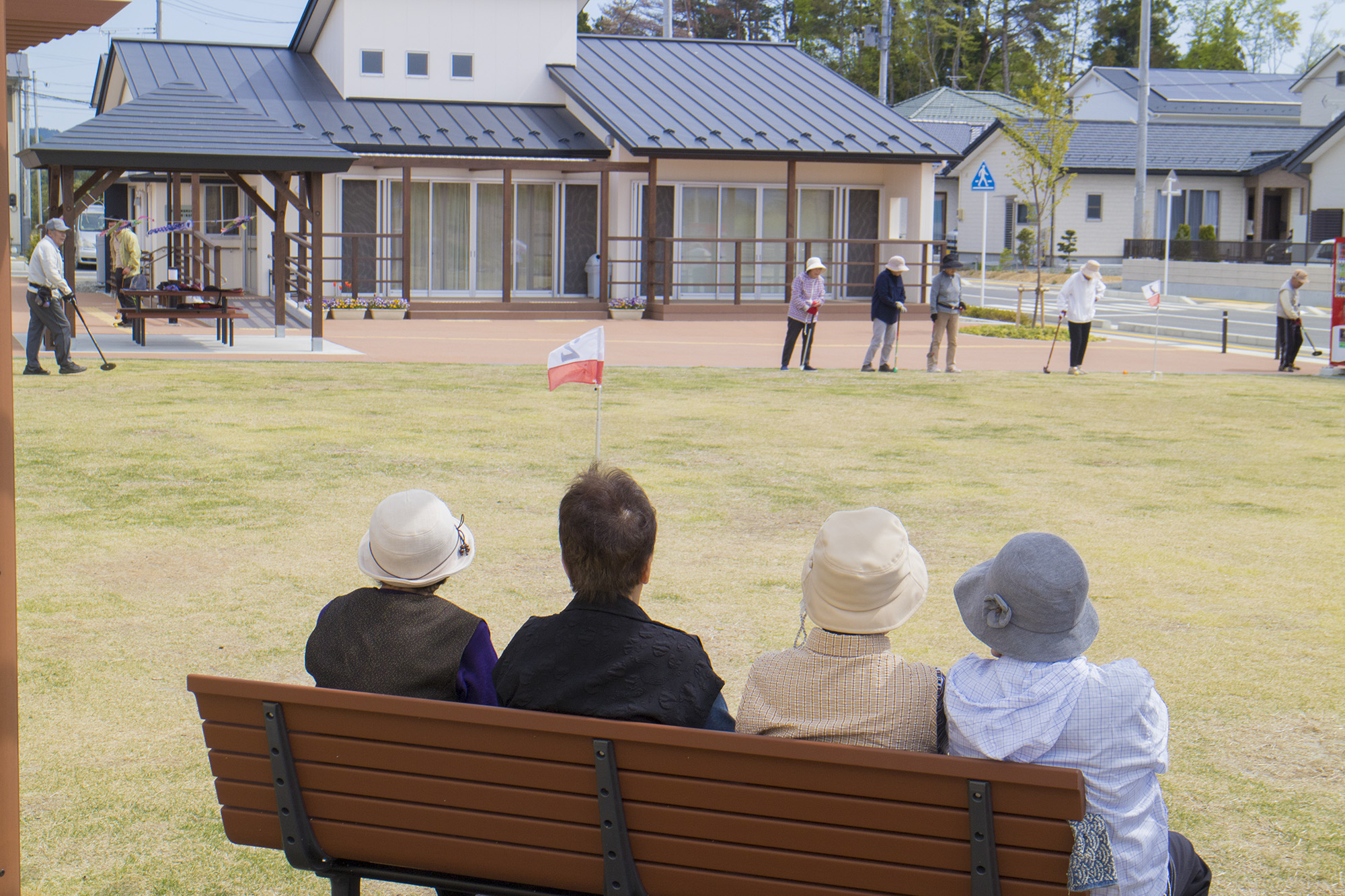 数人の高齢者がベンチに座って芝生の広場を眺めており、その前方では別の高齢者たちがグラウンドゴルフを楽しんでいる様子を写した写真