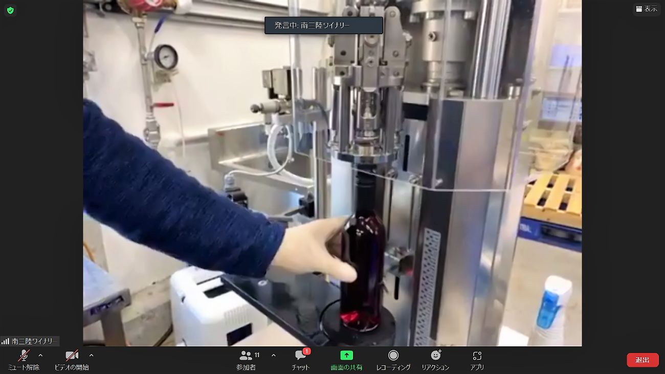 手袋をした人物がワインの瓶を専用の機械にセットしている瓶詰め作業の一部が映されているワイン製造現場の様子をZoomで映している写真