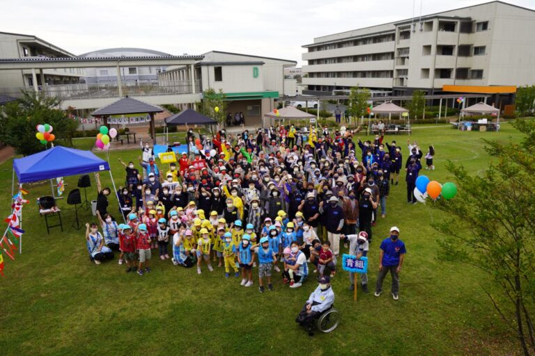 芝生広場に集まった子どもから高齢者までの多世代の参加者が色とりどりの衣装や帽子を身につけ、テントや風船などの装飾のもとで楽しげな集合写真