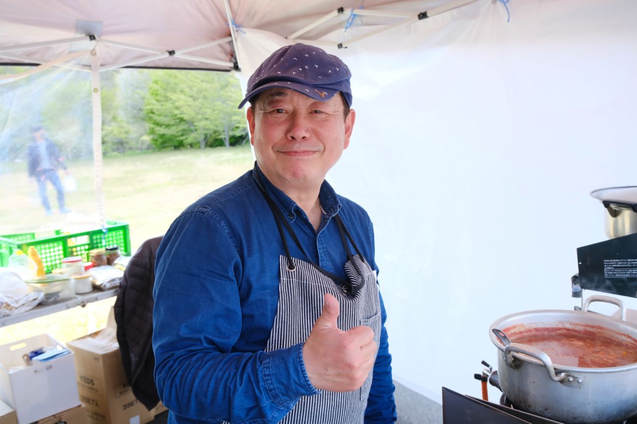 鍋で煮込まれている料理が見えるテントの中で、エプロンとキャップを着けた男性が、右手で親指を立てて笑顔を見せている写真