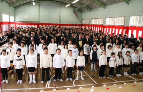紅白幕が飾られた体育館で、大勢の子どもたち関係者が集まり、整列して全員で校歌を歌っている閉校式典の様子の写真