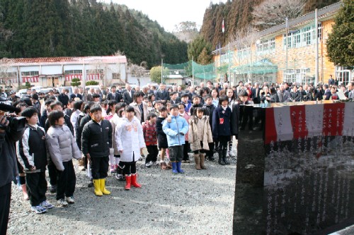 山々に囲まれた学校の校庭で、たくさんの子どもたちとその後ろに並ぶ大勢の関係者が、記念碑を向いて立っている様子の写真