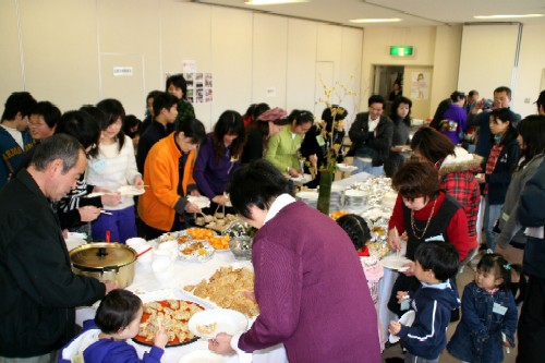 屋内の会場で、大人から子どもまで大勢の人々が、テーブルに並べられた料理を囲んで立食形式で食事を楽しんでいる国際交流フェアの写真