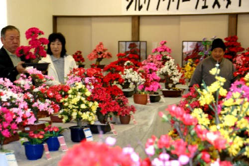 室内会場のテーブルの上に、ピンク、赤、白、黄色の鮮やかなシルクフラワーの鉢植えや盆栽が多数並べられ、数人の来場者が鑑賞している写真