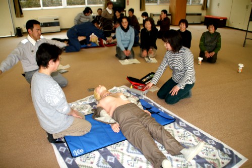 カーペット敷きの室内で、2つのグループに分かれた参加者たちが救命講習を受けており、手前のグループでは、消防署員の指導のもと参加者の女性2人が人形のモデルを使ってAEDの操作を体験している様子を他の参加者が座って見学している写真