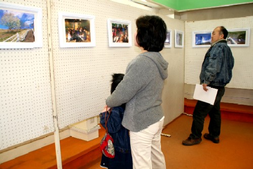 屋内壁面に展示された写真の数々を、小さい子どもを連れた女性や書類を手に持った男性が鑑賞している写真展の様子の写真