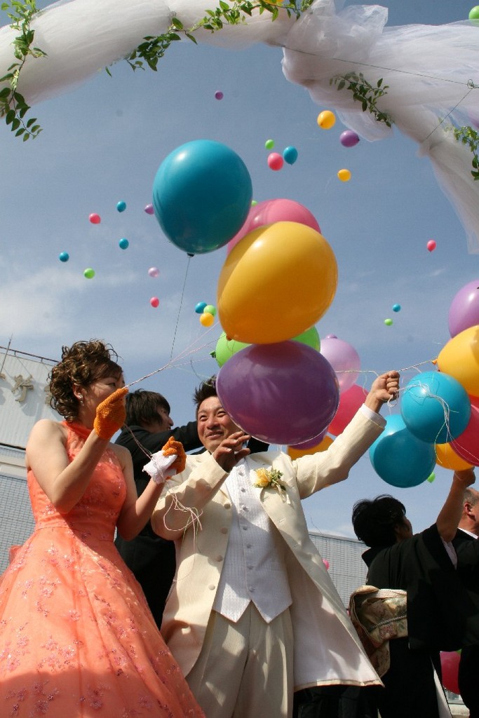 晴れた屋外で、白いチュールでできたアーチの下に立ったオレンジ色のドレスを着た新婦と、クリーム色のタキシードを着た新郎、礼服姿の参式者たちがカラフルな風船を手に持ち、空にたくさんの風船が舞い上がっているバルーンリリースの様子の写真