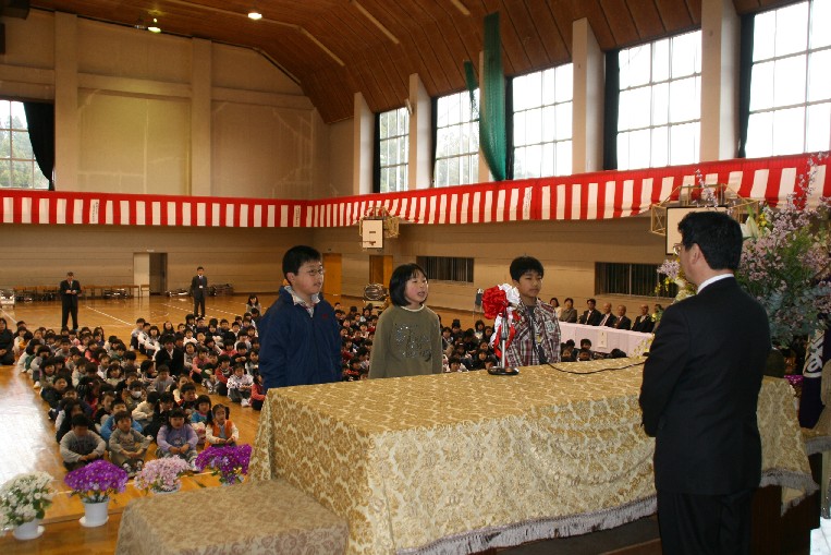紅白幕が張られた体育館に大勢の生徒が集まって整列して床に座り、来賓が席に座る中、壇上に上がった3人の児童が、演台に立ったスーツ姿の男性の前に並んで宣誓をしている様子の写真