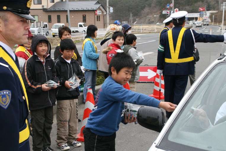 数人の警察官と一緒に黄色のタスキをかけた交通指導隊たちと袋詰めのわかめを持った数人の児童が駐車場に立って安全運転を呼びかける活動をしており、停車した車の運転手に男子児童がわかめの入った袋を手渡している様子の写真