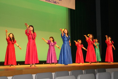 舞台の上で、赤色、紫色、青色、緑色の鮮やかなロングドレスを着用し、首飾りをつけた8人の女性たちが、手を上げてステップを踏む優雅なハワイアンダンスを披露している写真