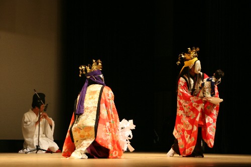 舞台上で、金色の冠と華やかな赤とオレンジ色の和装を身にまとい白い面をつけた二人の演者が、一人は背を向け跪き、もう一人は人形を抱いて立ち、左奥では和装の奏者が笛を奏でている伝統芸能の舞台の写真