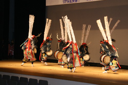 舞台上で色鮮やかな衣装を身に着け、白い羽の飾りが付いた頭飾りをかぶった出演者たちが、胴に太鼓をつけて踊りながら演奏している様子を写した、三人舞の写真