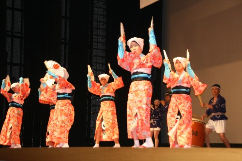 舞台上でオレンジの花柄の着物に黒地に青い帯、白い頭巾を身につけた踊り手たちが扇を手に腕を高く上げて踊っており、後方には太鼓を演奏する奏者が並ぶ、はねこ踊りの写真
