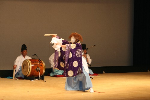 舞台の上で、白い面をつけ紫地に白菊の模様が入った装束を着た演者が、扇子をかざして舞を披露しており、その後ろには太鼓や弦楽器を演奏する伴奏者が床に座って演奏をしている、日本の伝統的な古典芸能の舞台の写真
