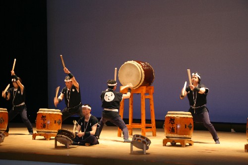 舞台上で揃いの衣装と鉢巻きを身に着けた5人の奏者たちが、力強いバチさばきで複数の太鼓を演奏している、和太鼓演奏の様子を写した写真