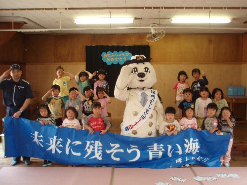 海上保安庁の制服を着た職員とマスコットキャラクターの「うーみん」、参加者の園児たちが並び、全員で敬礼のポーズをしながら、「未来に残そう青い海」と書かれた青い横断幕を広げて写っている記念写真