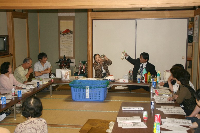 和室の部屋に、ロの字型に設置された長机に参加者の方々が座り、中央に青色のバスケットが置かれ、前方中央に座った女性と男性がペットボトルを持ち上げ、説明をしている様子の写真