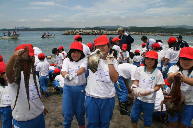 小舟が浮かび、防波ブロックが設置された海岸で、体操服に赤い帽子をかぶった子どもたちが地引き網に集まり、その手前では5人の子どもたちが、手に持った大きなタコや魚を嬉しそうにカメラに向かって見せている写真