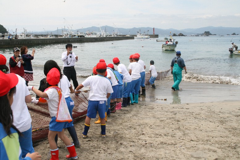 港に停泊する船や海上の小舟、遠くに山々が見える砂浜で、体操服に雨靴姿の子ども達が大きな網を海から引き上げる地引き網漁の体験をしている様子を写した写真
