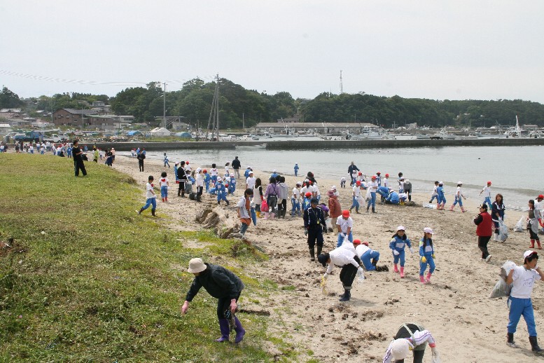 海沿いの砂浜と草地で、多くの子どもや大人の参加者が、清掃用具やゴミ袋を持ち、海岸に散乱したゴミを拾い集めている海岸清掃活動の様子の写