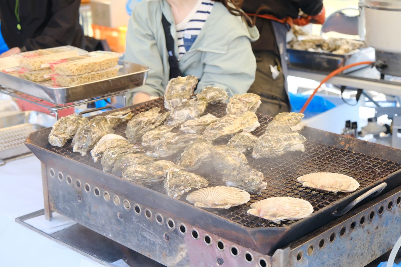 焼きそばのパックが置かれた後ろに女性スタッフが座っており、手前の網の上で牡蠣やホタテを焼いて湯気が出ている様子の写真