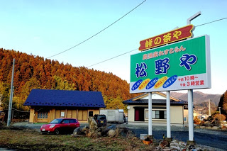 「峠の茶や 松乃や」と書かれた大きな看板が立っており、その奥に平屋建ての建物が見える農漁家レストラン・松野やの外観の写真
