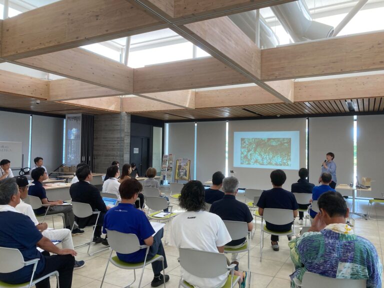 自然光が差し込む木造天井の会議室で、多くの人が前方のスクリーンを見ながら座ってプレゼンテーションを受けている様子の写真