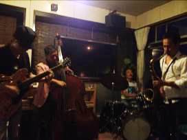 暗めの照明の室内で、4人の男性がギター、ウッドベース、ドラム、サックスを演奏している写真