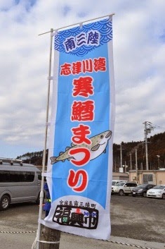 寒空の下、タラの魚のイラストとともに「南三陸 志津川湾 寒鱈まつり」と書かれたのぼり旗がポールに取り付けられはためいている写真