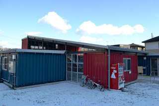 青と赤に塗られたコンテナで作られた建物の前に自動販売機が設置され、近くに自転車が二台停められ、地面には雪が積もった冬の風景の写真