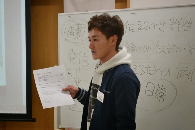 「情報」「発信」「日常」などの言葉が書かれているホワイトボードの前に立つ男性が、紙を手に持ちながら発表している写真