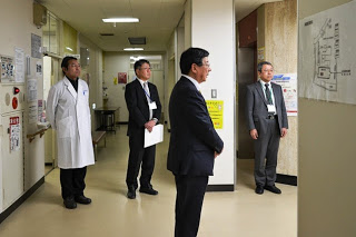 病院の廊下にスーツ姿の男性2人と白衣姿の男性1人が立ち、手前に立つ佐藤町長の話を聞いている写真