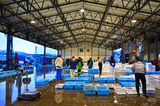 市場の屋内では多数の人々が白い発泡スチロールの箱や青い水槽の周囲に集まっている初セリ会場の様子の写真