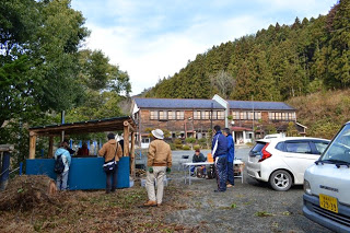 山あいの里山にある木造の旧校舎を背景に、屋外の作業小屋で数人の人々が作業をしている様子を写した、炭焼き体験の風景の写真