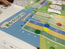 「みんなのまちづくりゲーム」と書かれたボードゲームの用紙の上に、緑、黄色、青、赤、白ののカラーチップやカードなどが置かれている写真