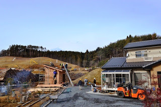 緩やかな丘と森に囲まれた高台の隣には住宅が建ち、重機が置かれた敷地内で複数の人が小屋を建てる作業をしている写真