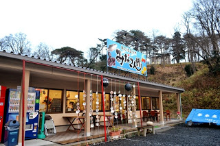 林に囲まれた小高い場所に、左側に自動販売機が設置された「みなさん館」と書かれた水色の看板のある平屋の店の外観写真