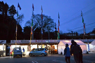 夕暮れ後の駐車場に沢山の旗が立てられている仮設店舗と車が並び、親子連れがお店に向かって歩いている写真