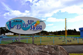 「平成の森しおかぜ球場」と書かれた大きな看板の向こうで、芝生のグラウンドに多くの人々が集まり活動している様子の写真
