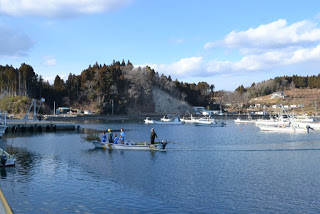 穏やかな晴天の下、南三陸の港で作業着を着た漁師たちが小型の漁船に乗って網を引いている様子の写真