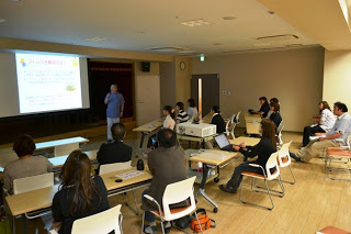 会議室で十数名の参加者が椅子に座って前方のスクリーンを見つめながら、男性講師の話を聞いている写真