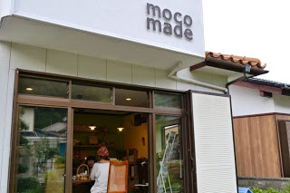 白い外壁に「moco made」と書かれた看板があり、ガラス戸の中で赤いバンダナを被った女性が作業している小さな店舗の写真