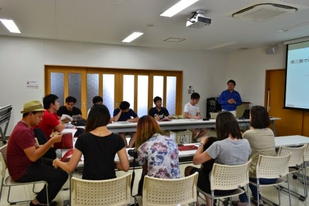 白壁の会議室で十数人がコの字型に設置された机を囲み、前方で男性がスライドを使いながら話をしている様子の写真