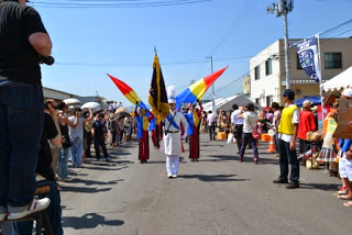 両脇に多くの観客が並んで見守っているなか、白い制服に身を包んだドラムメジャーを先頭に、お揃いの赤色の衣装を身にまとった創価ルネサンスバンガードのメンバーが、色とりどりの旗を掲げて堂々と行進している夏祭りのパレードの写真