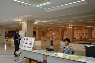 広い室内の展示会場で、入口近くの受付に座る人と、その近くに立っている人がいる様子が見える模型展の受付を写した写真