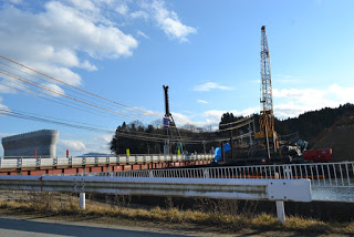 空は晴れて雲が少しあり、周囲には木々や丘が背景にある橋の上の工事現場で、クレーンやその他の建設機材があり、作業員が橋の建設または補修に関わっている様子が見られる写真