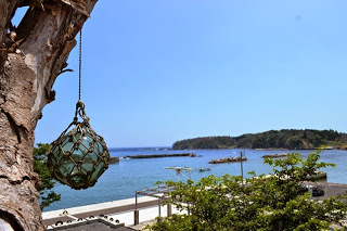 木に吊るされたガラス製の浮き玉越しに、青い海と防波堤、緑に覆われた小島が見える静かな漁港の風景の写真