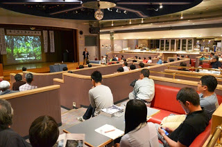 赤いソファ席が並ぶ広いホールで、スクリーンに映像が映し出され、来場者が静かに講演を聴いている会場全体の写真