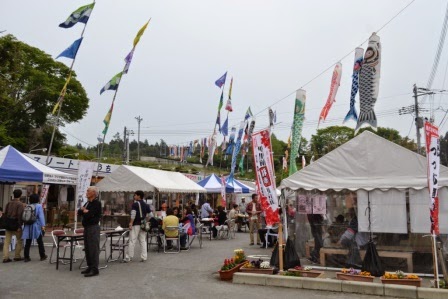 たくさんの鯉のぼりが空に掲げられた中で、テントの屋台が並び、会場に設置されたテーブルとイスに来場者が座って食事をしたり歩いたりしている祭り会場の写真